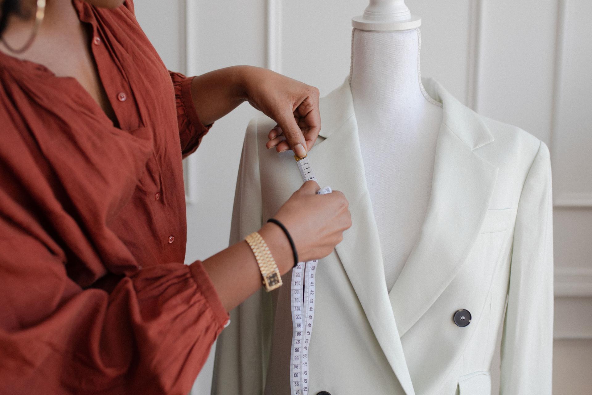 A black woman fashion designer measuring a white blazer on a dress form.